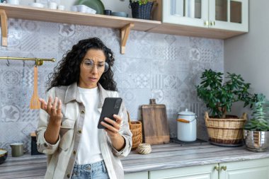 Depresyondaki üzgün İspanyol kadın akıllı telefondan kötü haberler okuyor, gözlüklü ve kıvırcık saçlı kadın mutfakta akıllı telefon kullanıyor..