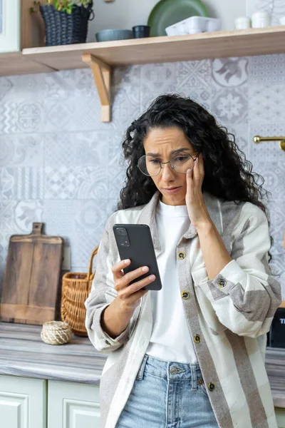 Dikey çekim, telefonda kötü haber okuyan üzgün kadın, kıvırcık saçlı ve gözlüklü İspanyol kadın akıllı telefon tutuyor..