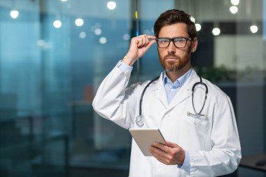 Sakallı olgun bir doktorun portresi. Gözlüklü bir adam kameraya bakıyor ve elinde tablet bir bilgisayar tutuyor. Doktor modern bir kliniğin ofisinde çalışıyor..