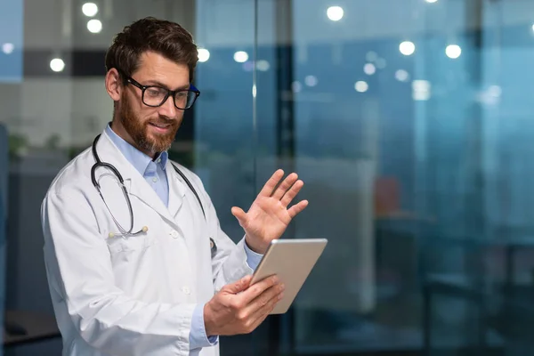 穿着医疗服的快乐的资深医生使用平板电脑与病人 留着胡子和眼镜的医生在诊所里进行视频通话和在线咨询 — 图库照片
