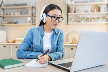 Online eğitim. Genç, güzel bir Asyalı kız beyaz kulaklıklarla ve dizüstü bilgisayarla evdeki masada oturuyor. Uzaktan çalışıyor. Bir ders, bir not defterine sınıf yazıyor..