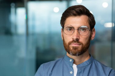 Ciddi ve düşünceli, sakallı, gömlekli ve gözlüklü, modern ofis binasında çalışmaya başlayan bir adamın yakın plan portresi..