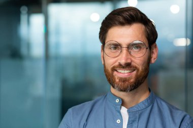 Sakallı ve gözlüklü başarılı ve neşeli bir iş adamı kameraya bakıyor ve gülümsüyor, modern ofisteki bir adamın yakın plan fotoğrafı..