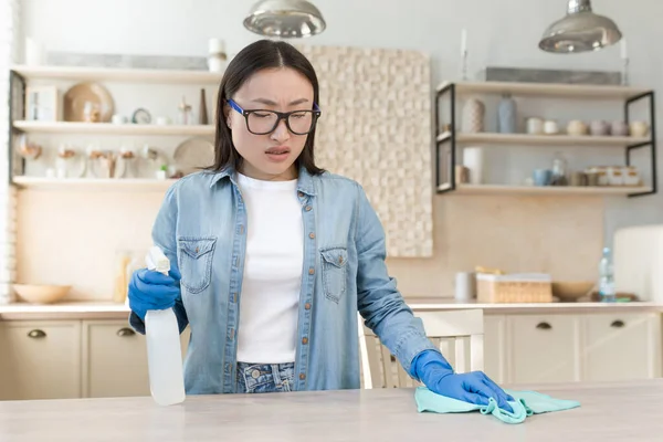 Quarantine means. A young beautiful Asian woman disinfects the house, wipes the table with a cloth in blue rubber gloves, sprays the product. Bends from dirt and dust.