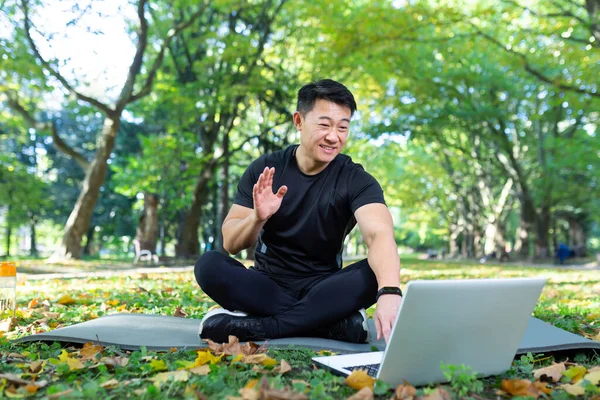 Fitness instructor works out online uses laptop in park sitting in lotus position, Asian athlete communicates with students remotely, video call training online remotely