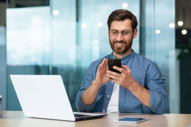 Başarılı ve mutlu sakallı iş adamı modern parlak ofiste çalışıyor, erkek patron dizüstü bilgisayar kullanıyor, iş sahibi sıradan gömlek giyiyor telefonu tutuyor, internet sayfalarını karıştırıyor