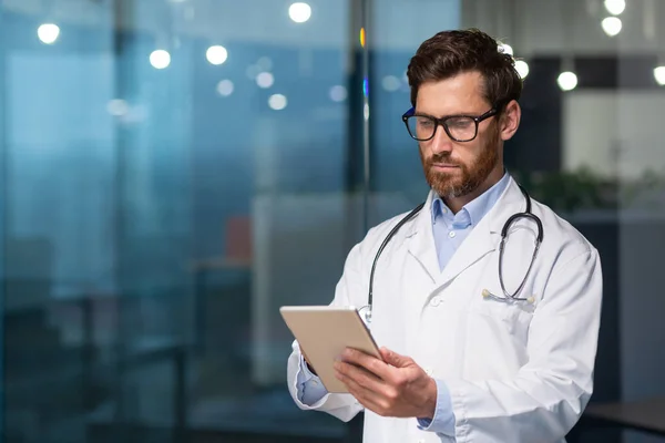 一位认真思考的医生从平板电脑上阅读医疗信息 一位身穿长袍的男子使用平板电脑 他可以集中注意力 输入信息 并查看病人的记录 — 图库照片