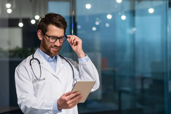 身披眼镜 身穿医疗服 面带笑容的资深医生 在现代诊所工作的人 正在阅读平板电脑发来的信息 — 图库照片