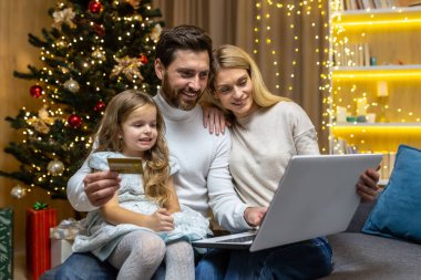 Noel ailesi, karı koca ve küçük kız Noel için evde online alışveriş yapıyor uzaktan kumandalı, güler yüzlü ve mutlu koca elinde banka kartı ve dizüstü bilgisayarla hediyeler seçiyor..