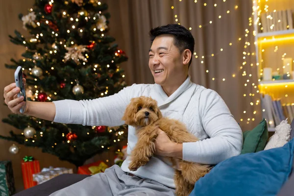 Christmas Eve Asian man with small pet dog talking on video call with friends, man sitting on sofa near Christmas tree celebrating new year waiting for friends to party.