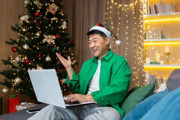 Cheerful man in christmas hat and green shirt talking with friends on video call, asian man celebrating christmas and new year at home sitting on sofa in living room with laptop.