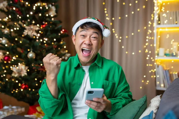 Portrait of successful man on New Years Eve, Asian man looking at camera and celebrating victory, holding phone received good news and online win, sitting on sofa near Christmas tree.
