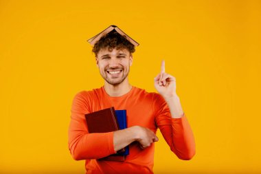 Kafasında kitapla gülümseyen genç ve yakışıklı bir adam, sarı arka plandaki sınavlara parmak kaldırarak hazırlanıyor. Yüksek kalite fotoğraf