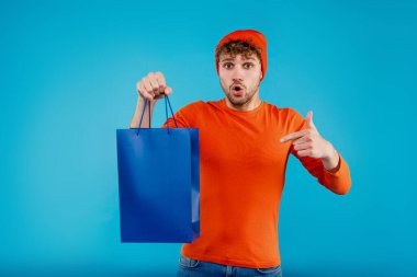 Genç, şaşırmış bir adam mavi arka planda alışveriş çantası tutuyor. Yüksek kalite fotoğraf