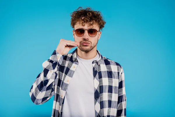 Young attractive man in printed shirt demonstrates gesture keep your mouth shut stands on blue background. High quality photo