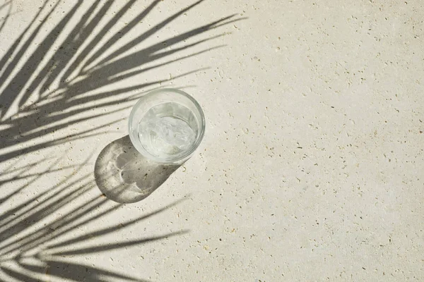 Trend glass of sparkling water from glass with palm leaf shadows on travertine stone background. Summer drink at sunlight. Minimal top view — Foto Stock