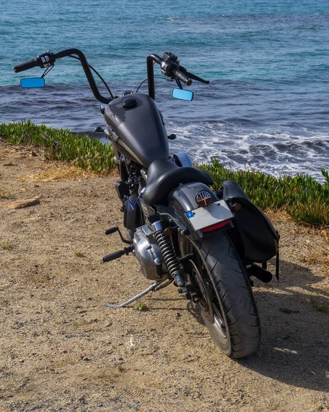 A black classic motorcycle stands on a cliff above the ocean on a sunny day