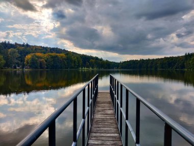 Göle giden dar iskele yukarıdaki bulutlu gökyüzünü yansıtıyor. Gölün etrafında bir orman var..