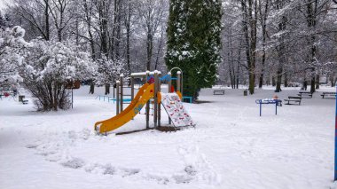 Kamu parkında çalılarla ve ağaçlarla çevrili taze beyaz karla kaplı eğimli bir çocuk kaydırağı..