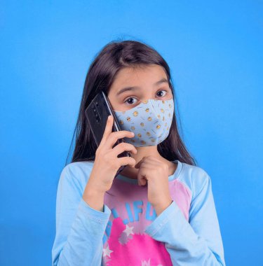 Koruyucu maskeli bir kız elinde cep telefonu, mavi arka planda izole edilmiş, kabarık saçlı 22 yaşında, renkli maskeli komik bir kız. Heyecan ve cazibe konsepti. Coronavirus mavi arka plan. Aptalca somurtmalar komik çılgın jestler. Komik ifade. 