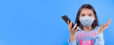 Koruyucu maskeli bir kız elinde cep telefonu, mavi arka planda izole edilmiş, kabarık saçlı 22 yaşında, renkli maskeli komik bir kız. Heyecan ve cazibe konsepti. Coronavirus mavi arka plan. Aptalca somurtmalar komik çılgın jestler. Komik ifade. 