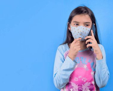 Koruyucu maskeli bir kız elinde cep telefonu, mavi arka planda izole edilmiş, kabarık saçlı 22 yaşında, renkli maskeli komik bir kız. Heyecan ve cazibe konsepti. Coronavirus mavi arka plan. Aptalca somurtmalar komik çılgın jestler. Komik ifade. 