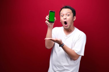 Şok Genç Asyalı adam kırmızı arka planda yeşil boş alanı işaret ediyor..