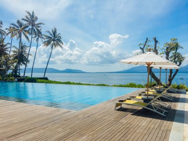 Şemsiyeli otel veya tatil beldesinde güzel tropikal yüzme havuzu, hindistan cevizi ağacı şezlongları, sonsuzluk havuzu manzaralı palmiye ağaçları, okyanus ve dağ arka planı.