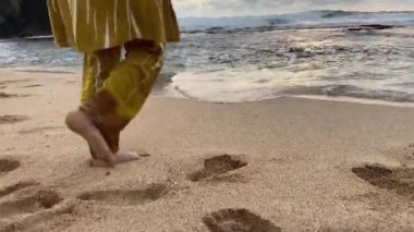 Slim female legs and feet walking along sea water waves on sandy beach.