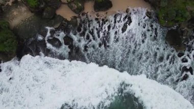 Okyanus dalgalarının mercan plajına çarpıp köpürmesini gösteren tepeden aşağı hava manzarası.