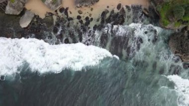 Okyanus dalgalarının mercan plajına çarpıp köpürmesini gösteren tepeden aşağı hava manzarası.