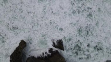 Okyanus dalgalarının mercan plajına çarpıp köpürmesini gösteren tepeden aşağı hava manzarası.