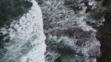 Okyanus dalgalarının mercan plajına çarpıp köpürmesini gösteren tepeden aşağı hava manzarası.