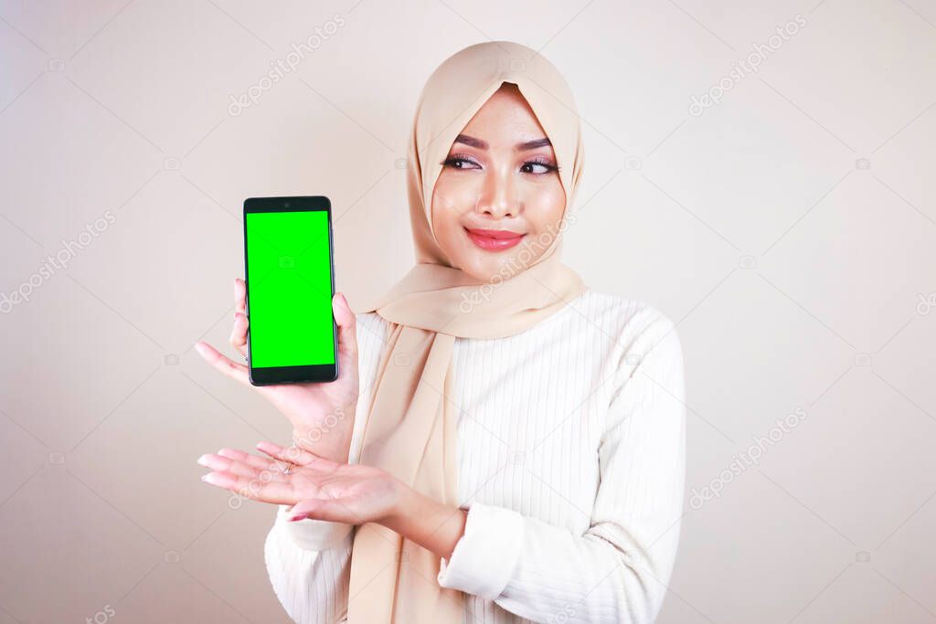 Portrait of cheerful young Muslim Asian woman pointing to cellphone with green screen