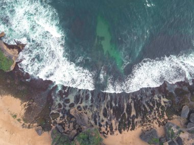 Okyanus dalgalarının mercan plajına çarpıp köpürmesini gösteren tepeden aşağı hava manzarası.