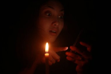 Asyalı bir kadın gecenin karanlığında cep telefonunu mum ışığının önüne doğrultuyor.