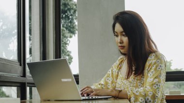 Young Asian woman working on laptop computer while sitting at the cafe, looking through documents