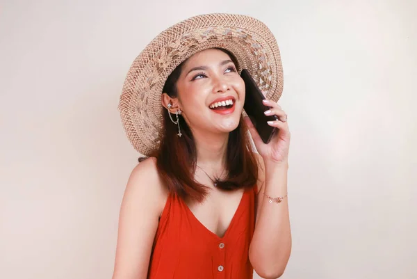 Happy Asian woman wearing a straw hat talking on a mobile phone and laughing, expressing positive emotions having a pleasant conversation