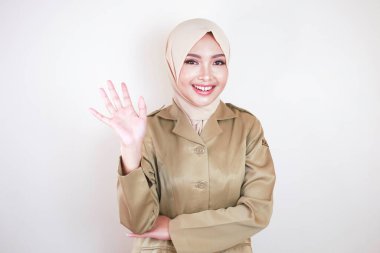 Müslüman sivil işçi kahverengi üniforma giyiyor ve tesettüre el hareketi yapıyor ve kameraya gülümsüyor.