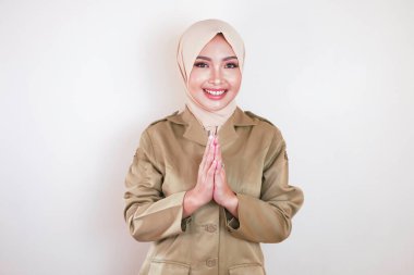 Müslüman sivil işçi kahverengi üniforma giyiyor ve tesettüre el hareketi yapıyor ve kameraya gülümsüyor.