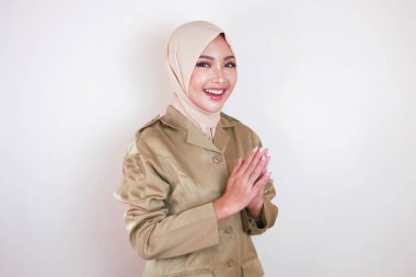 Müslüman sivil işçi kahverengi üniforma giyiyor ve tesettüre el hareketi yapıyor ve kameraya gülümsüyor.