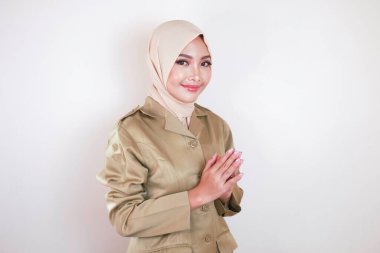 Müslüman sivil işçi kahverengi üniforma giyiyor ve tesettüre el hareketi yapıyor ve kameraya gülümsüyor.