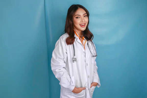 Confident relaxed female Asian doctor standing with hands in the pockets of her lab coat smiling quietly at the camera over blue background with copy space