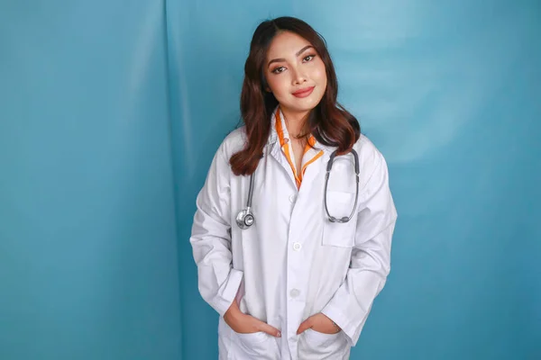 Confident relaxed female Asian doctor standing with hands in the pockets of her lab coat smiling quietly at the camera over blue background with copy space