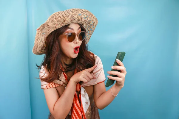 Young Asian woman pointing and shocking to her smartphone isolated by a blue background