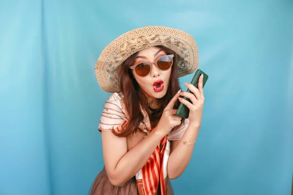 Young Asian woman pointing and shocking to her smartphone isolated by a blue background