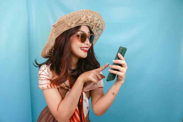 Genç Asyalı mutlu kadın mavi bir arka planla izole edilmiş akıllı telefonunu işaret ediyor ve gülümsüyor.