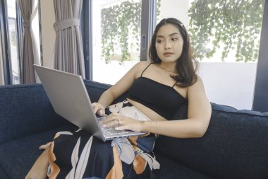 Oturma odasındaki rahat koltukta oturan Asyalı genç bir kadın dizüstü bilgisayarını fazla çalıştırmış ve yorgun görünüyor..