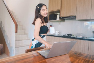 Genç Asyalı kadın zamanını yemek odasında uzaktan çalışarak bilgisayarında kupa taşırken çevrimiçi konferans yaparak geçiriyor..