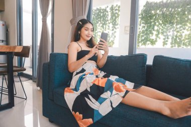 Genç Asyalı kadın boş zamanlarını oturma odasındaki rahat bir koltukta oturarak akıllı telefonundaki mobil uygulamadan mesaj göndererek geçiriyor..